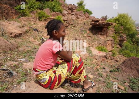 Side view of a serene daydreaming African villagegirl sitting on a rock amidst weeds and brushwood on a sunny day symbolizing differences in lifestyle Stock Photo