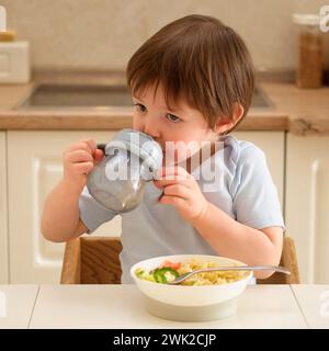 A child drinks from a non-watering cup while sitting at a table in a home kitchen. Cute little boy drinking water from a bowl in the kitchen at home. Stock Photo