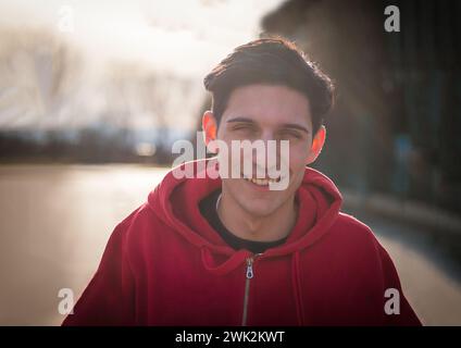A young man wearing a red hoodie smiles directly at the camera in this portrait photo. His expression is cheerful and inviting, exuding a sense of friendliness and approachability. Stock Photo
