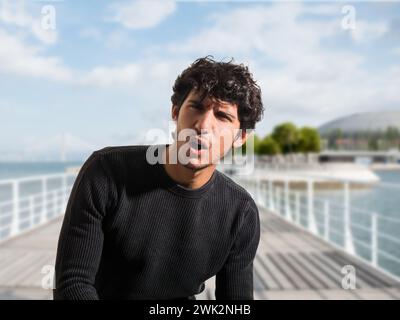 A man standing on a pier, looking surprised or hurting from pain, with his mouth open and eyebrows raised. The background shows water and boats. Stock Photo