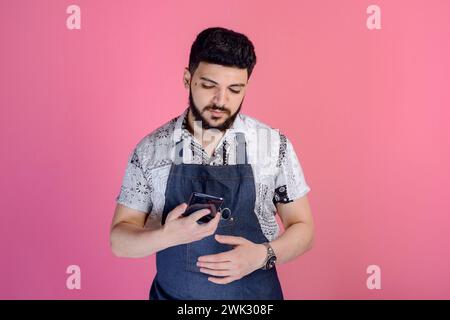 Brunette man wearing uniform, apron, holding smartphone, using, scrolling, texting. Stock Photo
