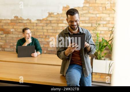 In a contemporary office setting, two focused colleagues engage in their work with tablet and laptop, collaborating and sharing ideas in a bright and Stock Photo