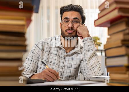 Exhausted Indian businessman reviewing documents reports while using calculator working hard in home office. Stressed alone freelancer touching head in despair surrounded by stacks of books on table. Stock Photo