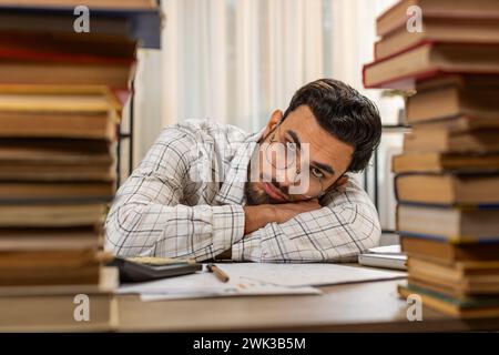 Exhausted Arabian businessman reviewing documents reports while using calculator working hard in home office. Stressed tired freelancer touching head in despair surrounded by stacks of books on table Stock Photo