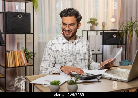 Focused young Indian office worker working with schedules and financial graphics while using calculator sitting at home office table. Happy businessman freelancer compare and analyze graphs charts Stock Photo