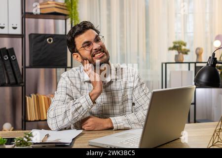 Indian businessman freelancer working on laptop and feeling toothache touching mouth with hand, painful expression because of dental illness. Caries, pulpitis, gum disease, dental disease concepts. Stock Photo