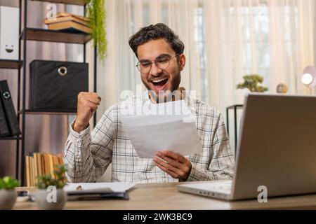Happy Indian young businessman manager open envelope letter, reading it. Career growth advance promotion, bank loan approval, successful admission to monetary award, long-awaited invitation great news Stock Photo