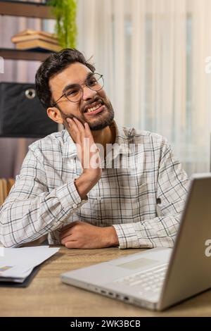 Indian businessman freelancer working on laptop, feeling toothache touching mouth with hand, painful expression because of dental illness. Caries pulpitis gum disease dental disease concepts. Vertical Stock Photo