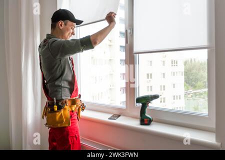 Maintenance man is fixing windows in a living room using a skrewdriver. He is wearing glasses, yellow gloves and a black apron Stock Photo