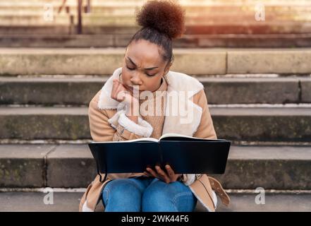 woman student e-learning distance training course study working on stairs in the city. African Ethnicity young woman watching online education webinar Stock Photo
