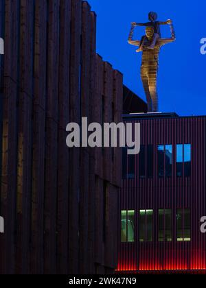 A striking illuminated sculpture of a human figure stands atop a modern building with vertical wood cladding. The photograph captures the contrast bet Stock Photo