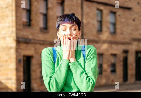 a woman in a green shirt shocked from news.  Holding hands on her mouth. Standing alone outside. Old buildings behind. Stock Photo