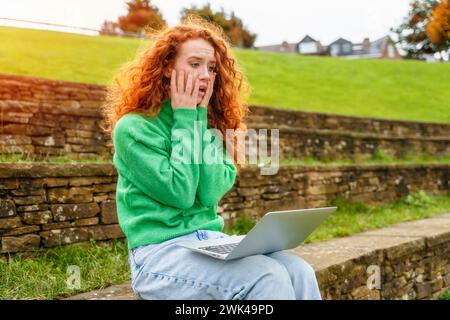 a stunned student redhead  woman working, studying, communicating on a laptop in a green public park of the university campus in the city center Stock Photo