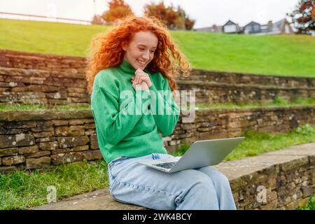 Portrait of beautiful happy young woman graduate with curly red hair reading e-mail on the laptop and excited  about news regarding her job applicatio Stock Photo