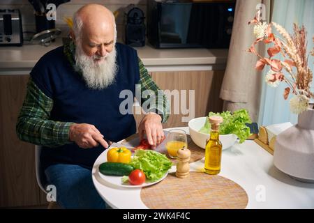 Old man at the kitchen table cuts vegetables for salad Stock Photo