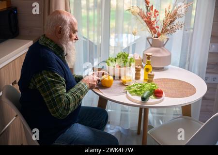 Gray-bearded man at the kitchen table cuts vegetables for salad Stock Photo