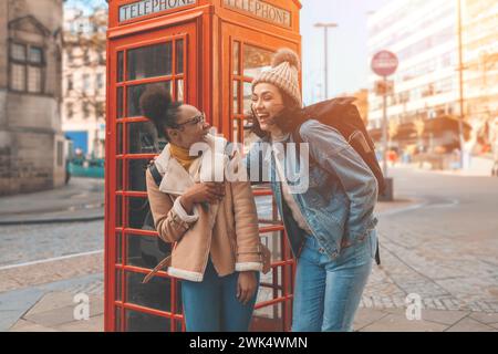 two friends, girlfriends and women using a mobile phone,  camera and taking selfie against a red phonebox in the city of England. Travel Lifestyle con Stock Photo