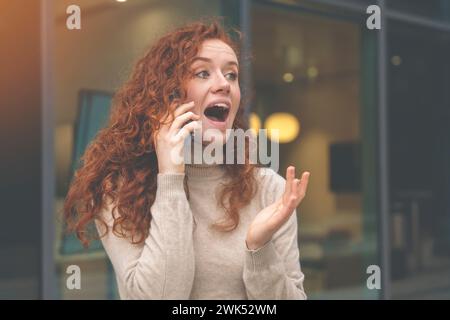 happy young woman graduate with curly red hair talking on the mobile and excited  about great  news regarding her job application Stock Photo