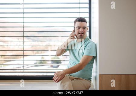Portrait of young man with Down syndrome sitting by window, making phonecall with smartphone. Stock Photo