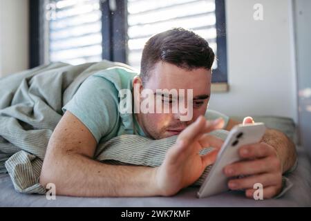 Young man with down syndrome lying in his bed, looking at smartphone in the morning. Morning routine for man with disability. Stock Photo