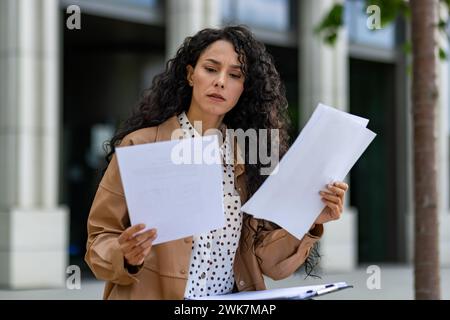 Professional young woman with curly hair scrutinizes papers during a lunch break by her office, embodying business acumen. Stock Photo