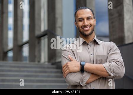 Portrait of a smiling young Muslim businessman in a shirt standing near an office building, smiling and confidently looking at the camera with his arms crossed on his chest. Stock Photo