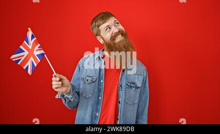 Handsome young redhead man with beard, oozing confidence, standing isolated over red background, holding high upside the great britain flag, smiling i Stock Photo