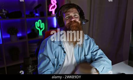 Staunch young redhead man streamer, seriously immersed in digital gaming world, donning headphones in the dimly lit gaming room Stock Photo