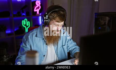Staunch young redhead man streamer, seriously immersed in digital gaming world, donning headphones in the dimly lit gaming room Stock Photo