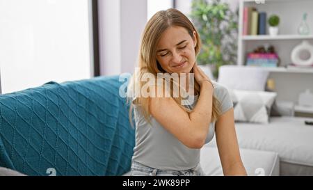 Exhausted young blonde woman sitting on sofa suffering from intense cervical pain indoors at home Stock Photo