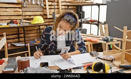 Young, beautiful hispanic woman carpenter counts dollars, taking business notes during woodwork session in indoor workshop Stock Photo