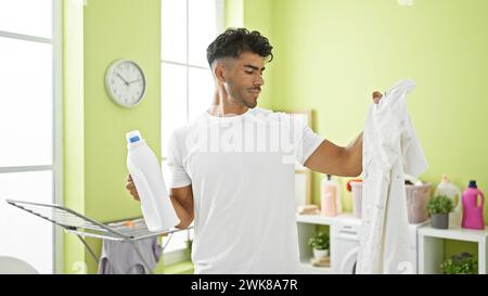 Handsome hispanic man doing laundry in a bright room, holding detergent and inspecting a white shirt. Stock Photo