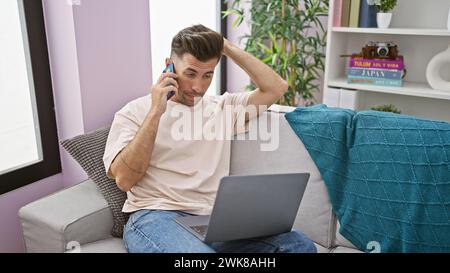 Stressed out young hispanic man, sitting on the sofa at home, engrossed in a tense phone conversation, working seriously on his laptop, totally focuse Stock Photo