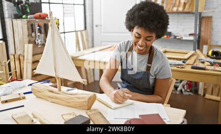 A smiling african american woman carpenter writing in a notebook in a woodwork studio workshop. Stock Photo