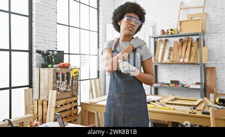 Black woman in safety glasses at a carpentry workshop holds shoulder implying injury indoors. Stock Photo