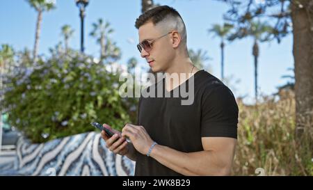 Handsome hispanic man using smartphone in a city park, surrounded by greenery and palm trees. Stock Photo