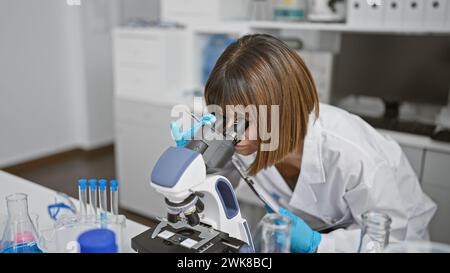 Relaxed yet focused, a young, beautiful hispanic woman scientist working diligently indoors at the lab, using a microscope. she's deeply engrossed in Stock Photo