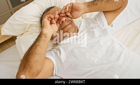 A mature hispanic man in a white shirt lying on a bed indoors, looking tired as he rubs his eyes. Stock Photo