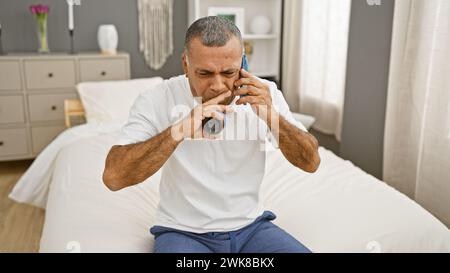 Middle-aged hispanic man using inhaler for asthma sitting on bed in a bedroom. Stock Photo