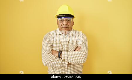 Confident middle-aged man with arms crossed wearing safety helmet and glasses against a yellow background. Stock Photo
