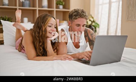 A smiling couple lounges in a cozy bedroom, browsing on a laptop together, evoking themes of love, togetherness, and domestic life. Stock Photo