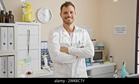 Handsome bearded young man in a lab coat stands confidently with arms crossed at a hospital clinic. Stock Photo