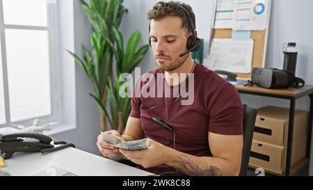 Hispanic man with a beard and tattoos counts money while wearing a headset in an office setting. Stock Photo