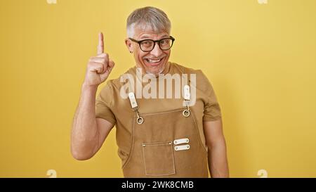 Cheerful middle-aged man with glasses and grey hair wearing suspenders, pointing up, isolated against a yellow background. Stock Photo