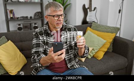 Middle-aged man checking medication bottle while using smartphone in living room. Stock Photo