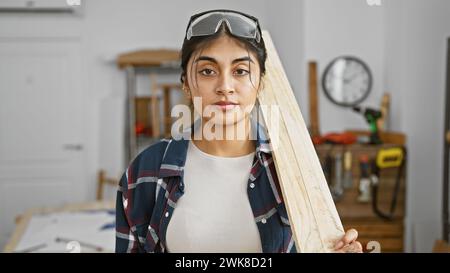 South asian woman with long hair wearing safety glasses in a woodworking workshop holding a plank of wood. Stock Photo