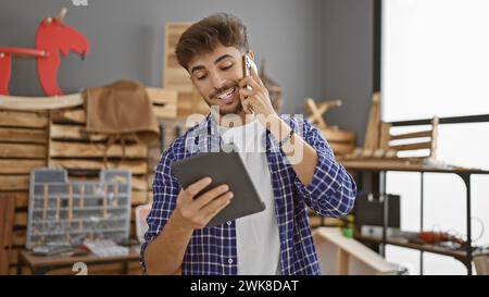 Handsome young arab man carpenter talking on smartphone, beaming with smiling face, engrossed in woodworking on touchpad indoors at his carpentry work Stock Photo