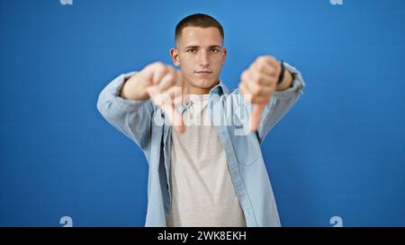 Hispanic man giving thumbs down gesture against a solid blue background, expressing disapproval Stock Photo