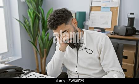 Tired hispanic man with beard using headset at office desk looking stressed Stock Photo
