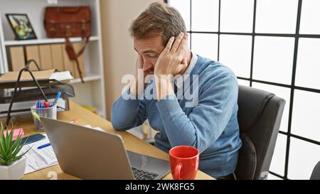 A stressed man with a beard sits in an office, touching his temple, in front of a laptop, showcasing a moment of frustration or fatigue. Stock Photo